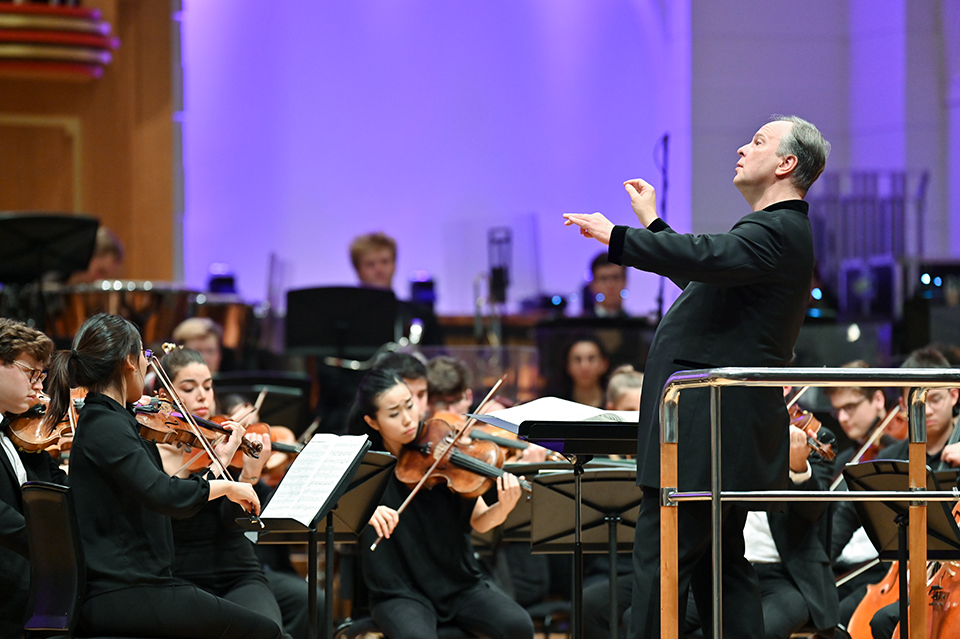A man dressed in black with his hands in the air conducting an orchestra.test